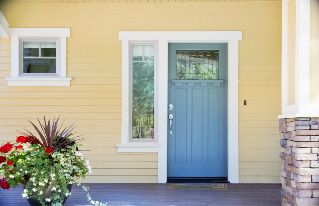 A front entrance of a home with a blue door, yellow siding, and a flowerpot in daytime.