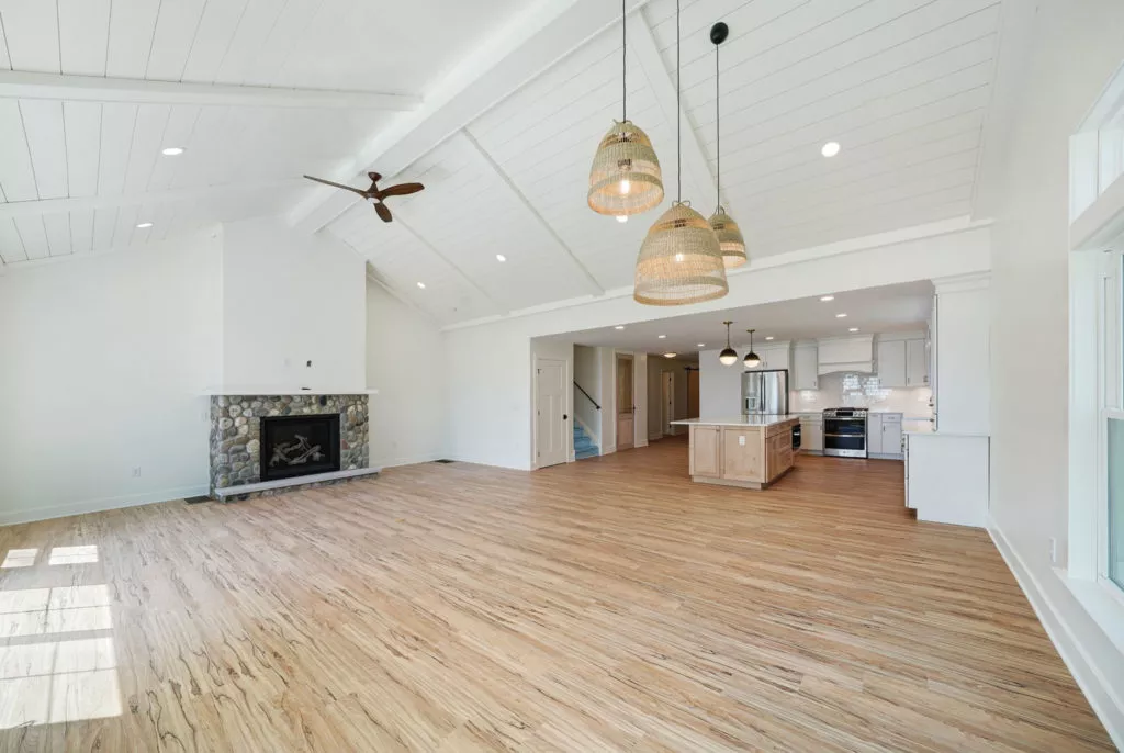 Inside a residential, two-story house's living room. A-frame ceiling and fireplace. Open concept kitchen with large kitchen island in background.