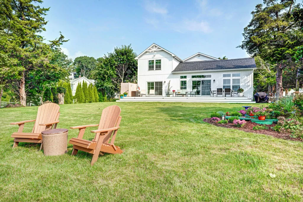 The back of a white, two-story house with large backyard, wooden chairs and deck.