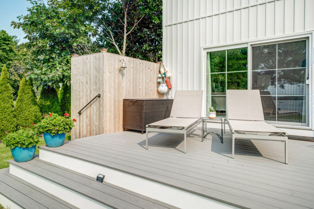 A gray porch with a tall wooden fence, flowers, and two lawn chairs.