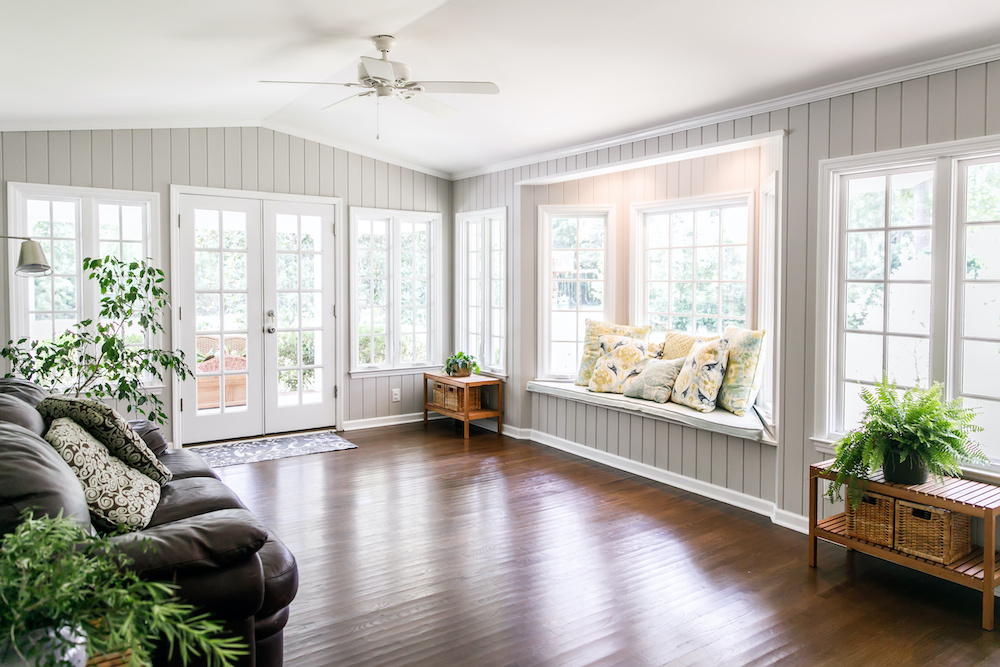 Large and open living room den sun room with windows on two sides and lots of natural light flowing in. There is a window seat on one side and a leather couch and plant on the other.