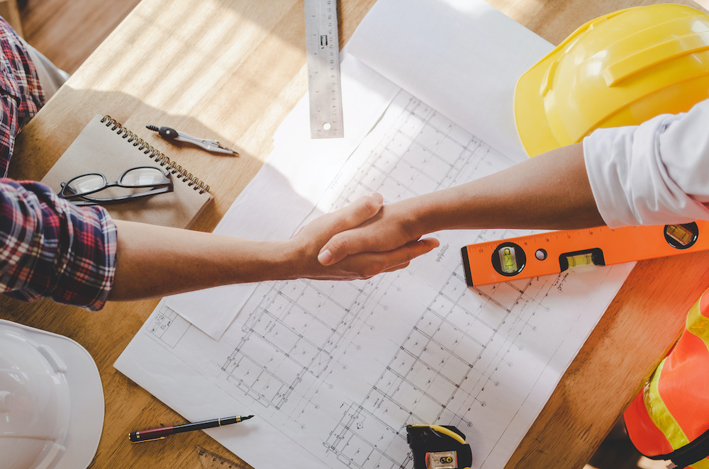 two people shaking hands over a table filled with blueprints and a hard hat